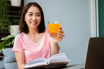 Mid-age Asian woman reading a book and hold orange juice glass in the home. Concept of  Health care And eating for healthy