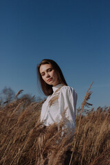 Smiling young woman in white blouse standing in field of dry pampas grass in front of sky and sun. Urban style and street fashion. Girl in casual outfit looking at camera. Golden hour