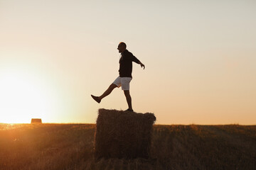 handsome young man in black sweatshirt and white short, in summer field outdoor, having fun on hay bale, haystack on sunset