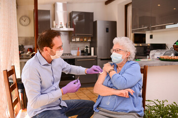 elderly senior woman with surgical mask having covid 19 vaccine injection by doctor male nurse at home