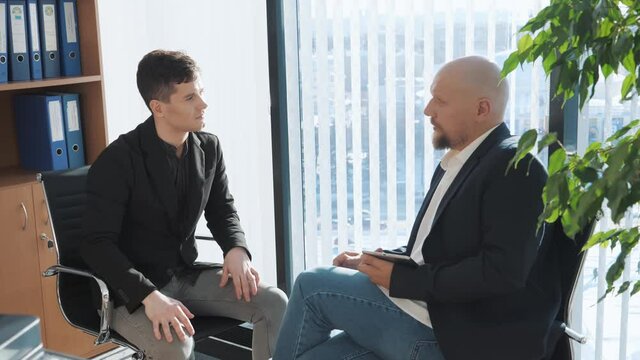 Portrait of two businessman sitting and chatting with office background. Casual looking men in suits discussing oportunities and upcoming projects. Concept of interview, deal.