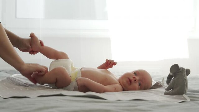Mother with baby boy doing exercise simple exercises to make baby bones and muscles stronger, newborn infant lying on bed against the window. High quality 4k footage