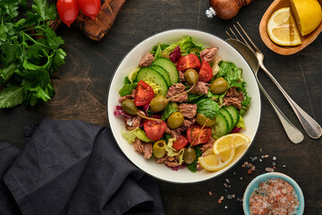 Tuna salad with fresh vegetables, olives, capers and lemon served in bowl on old wooden background. Top view with copy space.