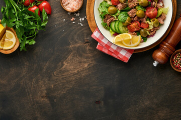 Tuna salad with fresh vegetables, olives, capers and lemon served in bowl on old wooden background. Top view with copy space.