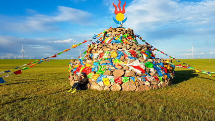 A woman in a beanie sitting on a vast pasture under a heap of stones (aobao) with colorful prayer...