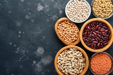 Legumes, red lentils, chickpea, red and white beans assortment in different bowls on dark black table. Diet for weight loss and fight against cholesterol concept. Top view with copy space.