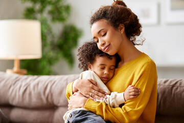 Loving afro american mom cuddling with little kid son while relaxing on sofa