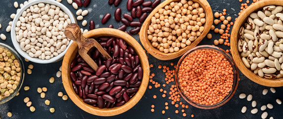 Legumes, red lentils, chickpea, red and white beans assortment in different bowls on dark black table. Diet for weight loss and fight against cholesterol concept. Top view with copy space.