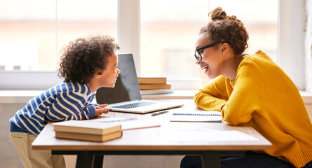 Young happy afro american woman mom talking with little kid son while working on laptop online at home
