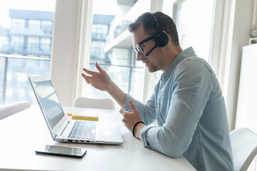 Side view at young male employee wearing headset talking online with a colleagues or customers, a...
