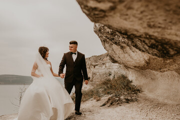 bride in white dress long veil and groom in suit walk in summer fall on mountain above river