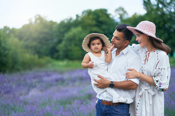 Happy family young couple posing with kid in lavender field. Little baby girl wearing straw hat sitting on hands of loving dad, looking at camera outdoors, aromatic flowers. Family, nature concept.