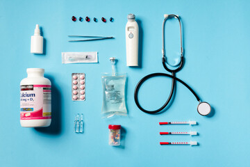 Overhead view of a medical instruments consisting of a jar of calcium, syringes, thermometer, stethoscope, tweezers lying on a blue background