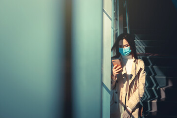 Girl with medical mask is using her smartphone in a modern building during pandemic of Coronavirus Covid-19