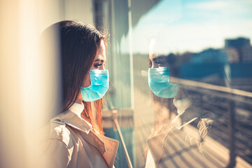 Girl with medical mask is looking out of the window during pandemic of Coronavirus Covid-19