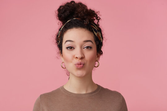 Portrait of charming girl with dark curly hair bun. Wearing headband, earrings and brown sweater. Has make up. Purses lips in a kiss. Watching at the camera isolated over pastel pink background