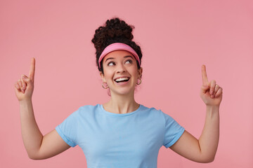 Playful woman, beautiful girl with dark curly hair bun. Wearing pink visor, earrings and blue t-shirt. Has make up. Pointing fingers and watching up at copy space, isolated over pastel pink background
