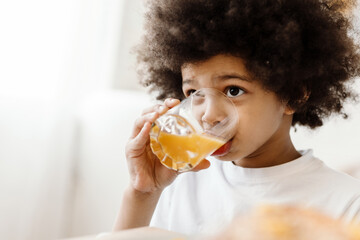 Black curly boy looking forward while drinking juice indoors - obrazy, fototapety, plakaty