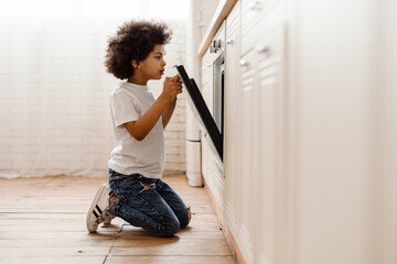 Black curly boy opening oven while sitting on floor in home kitchen