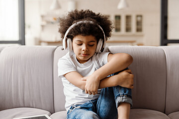 Black curly boy listening music with headphones while sitting on sofa