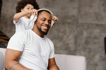 Black father and son smiling and using headphones while sitting on sofa