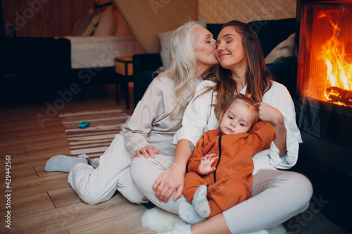 Grandmother, Mother and child kissing and playing on sofa near fireplace. Mom and baby. Parent with daughter and grandson little kid relaxing at home. Family having fun together. Mother's day
