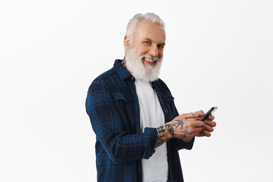 Happy stylish senior man using mobile phone, messaging or browsing internet on smartphone and looking at camera with cheerful smile, standing over white background
