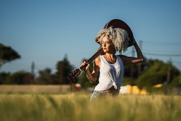 Chica guapa tatuada con pelo muy rizado posando en un campo de trigo con una guitarra acsutica