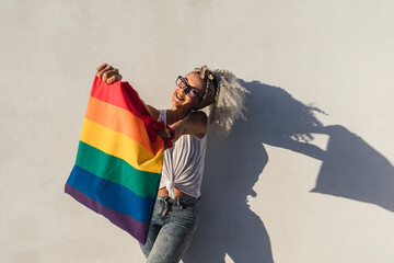 Chica delgada con pelo muy rizado delante de pared blanca posando feliz ocn una bolsa con la bandera de arco iris