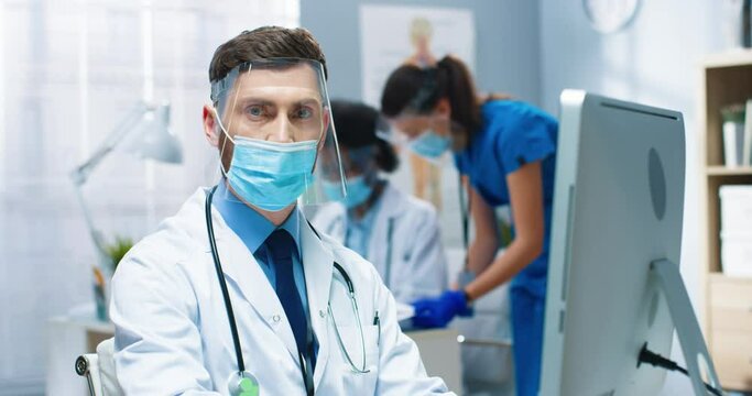 Close up portrait of handsome serious Caucasian male healthcare worker in mask and face shield working in hospital lab during coronavirus browsing on computer and looking at camera, clinic concept