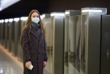A woman in a medical face mask is waiting for a train and holding a smartphone.