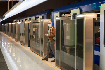 Man in a medical face mask is holding a phone while leaving a modern subway car.