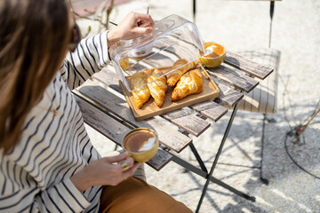 Young woman sitting at a table in the garden and having a breakfast. Eating croissant with coffee. French style. Spend time outdoors in the sun.