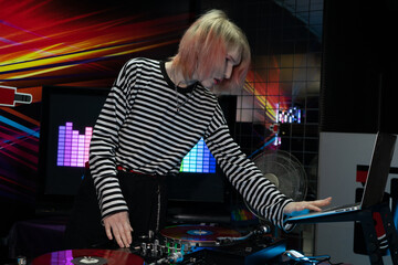 A transgender DJ tunes a song on a laptop, in front of him is a DJ equipment with two red records,...