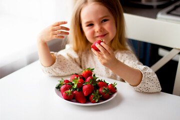 Cute little girl eating fresh strawberry in the kitchen. Healthy vitamin snack for kids. Ripe fresh berries. Harvest season. Natural vitamins .