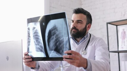 Young male doctor is looking at chest x-ray while sitting in medical office. Specialist examining x-ray picture and holding it in his hands, taking notes in medical form during working day in hospital - Powered by Adobe
