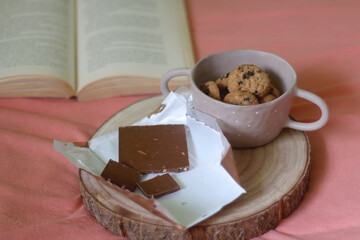 Chocolate bar, bowl of chocolate chip cookies and open book on a bed. Selective focus.
