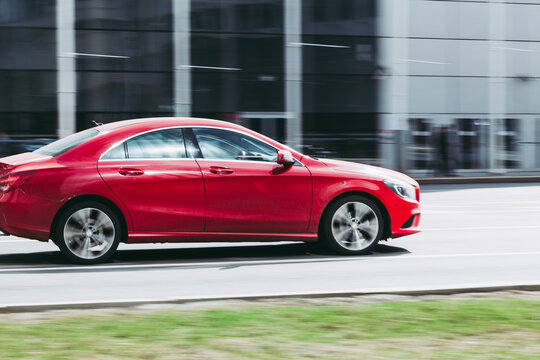 Side View Rolling Shot With Red Car In Motion. Mercedes CLA-Class Driving Along The Street In City With Blurred Background