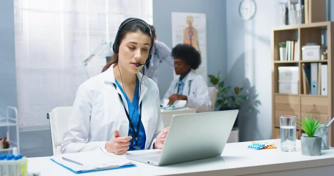 Caucasian young pretty female doctor having online video call consultation on laptop speaking in headset with patient sitting at table in hospital cabinet. Healthcare, Portrait, medical center concept