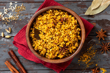 Cooked spicy rice in a wooden bowl and various spices on a dark background. Rice with cumin, turmeric, star anise and other spices. Indian food.