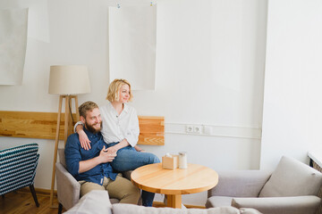 Fototapeta premium Romantic and happy caucasian couple in casual clothes sitting together in an armchair in cozy light space. Love, relationships, romance, happiness concept. Man and woman holding hands and smiling.