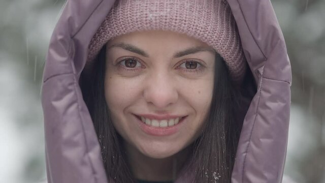 Close-up face of confident beautiful brunette woman with brown eyes and toothy smile looking at camera as snow falling outdoors. Headshot of happy carefree Caucasian tourist on winter resort