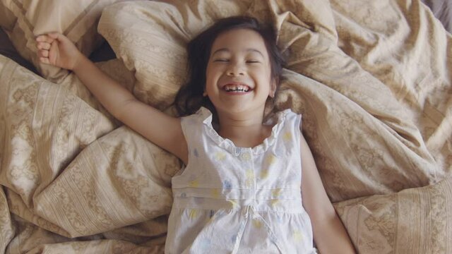 Portrait of adorable asian toddler with white skin in pyjamas who is acting on camera in the bed in the morning with sunlight shining through window. Her eye contact looking expresses joyfulness.

