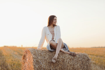 Naklejka na ściany i meble Young woman with long hair, wearing jeans skirt, light shirt and straw bag in hand, sitting on bale on field in summer. Female portrait in natural rural scene. Environmental eco tourism concept.
