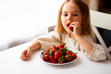Cute little girl eating fresh strawberry in the kitchen. Healthy vitamin snack for kids. Ripe fresh berries. Harvest season. Natural vitamins .