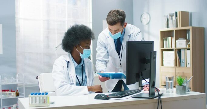 Mixed-race male and female doctors in medical masks speaking discussing analysis results working in hospital. African American young woman physician talking to Caucasian coworker. Healthcare concept