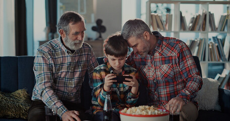 Three family people of father and son with elderly grandpa using mobile phone playing online videogame racing exciting together supporting the child.