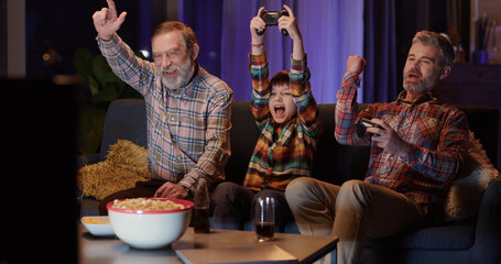 Multi-generational caucasian family of three playing console computer online video games together and eating popcorn snacks. Home entertainment. © Fractal Pictures