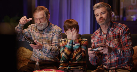 Multi-generational caucasian family of three playing console computer online video games together and eating popcorn snacks. Home entertainment.