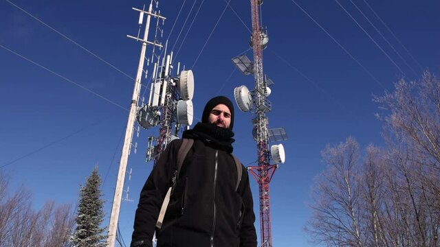 A tilting up video showing a passionate documentarian standing near tall 4G and 5G telecommunication towers, surrounded by pine trees.